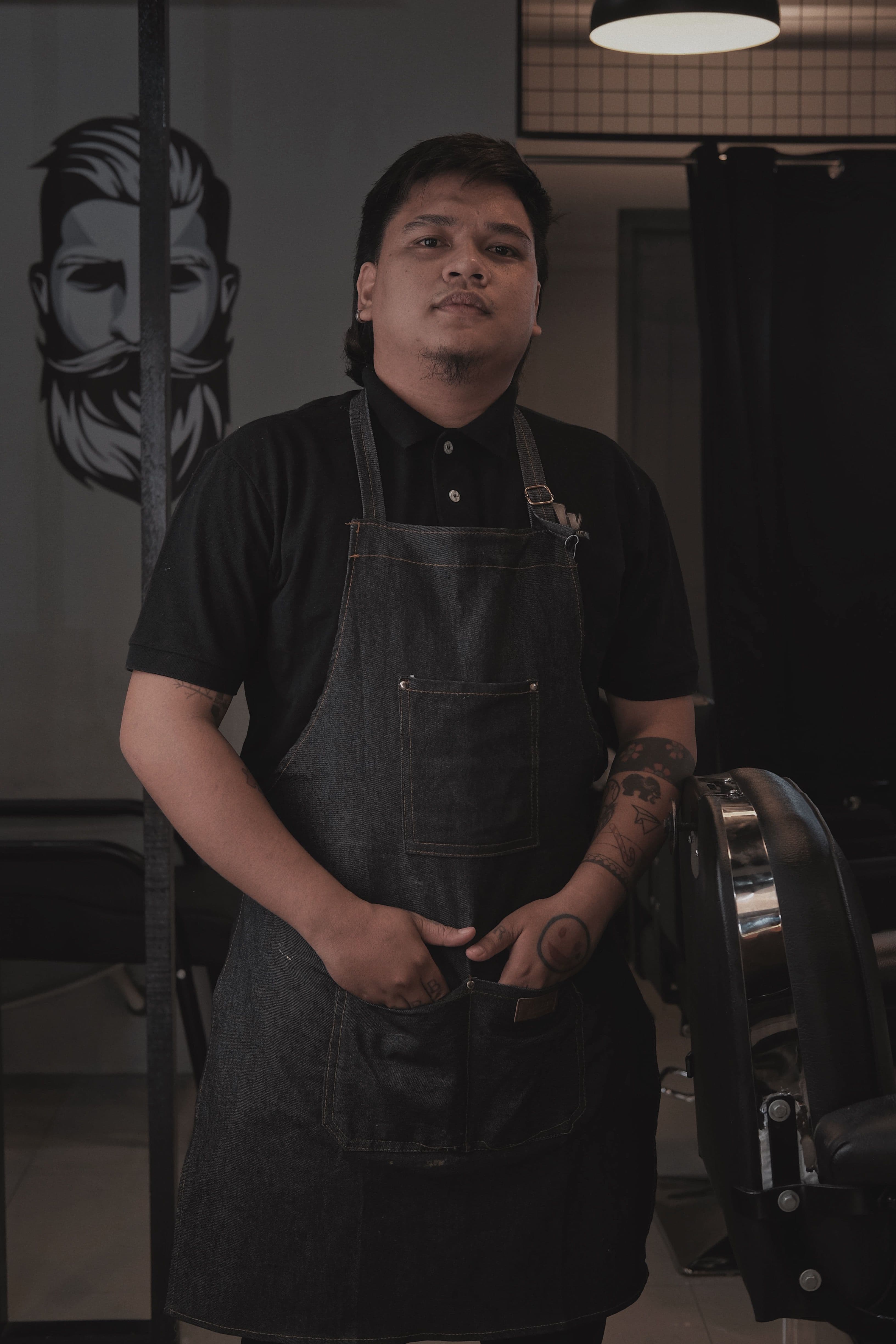 Portrait of barber Krit in a black polo and denim apron, standing next to a barber chair