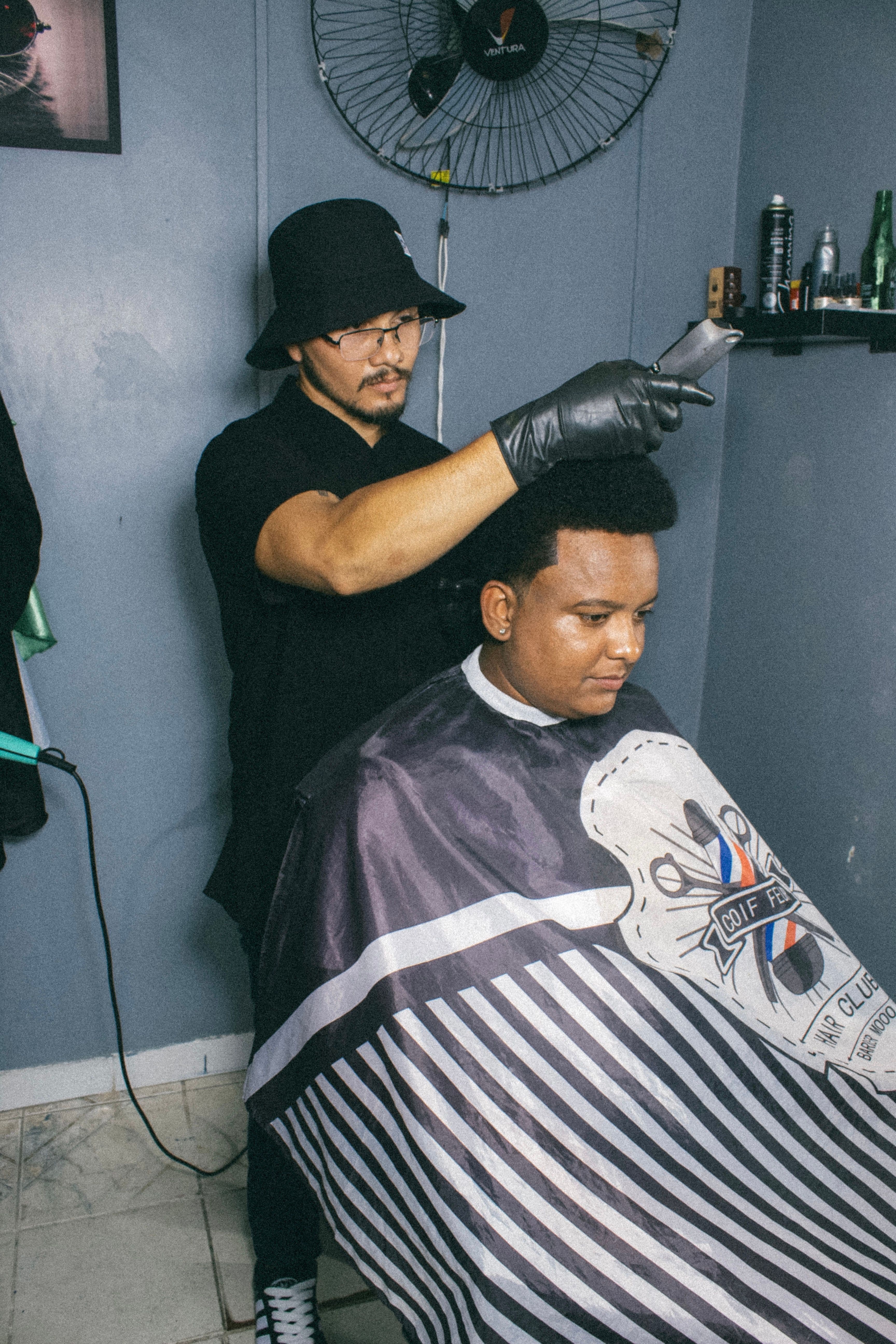 Barber Ton in a bucket hat and glasses fading a client’s hair with clippers