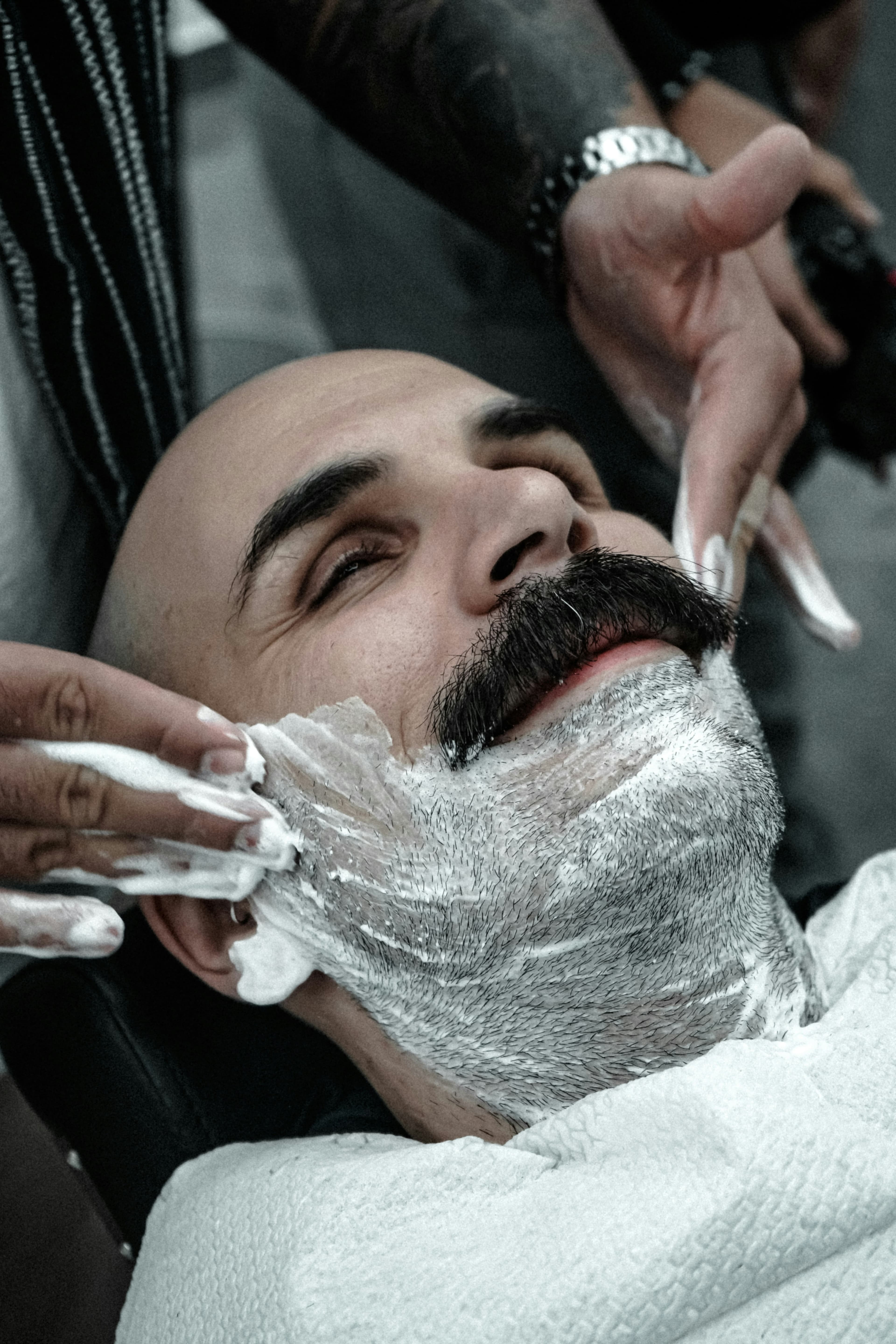 Client with a moustache reclining while shaving cream is applied before a razor shave